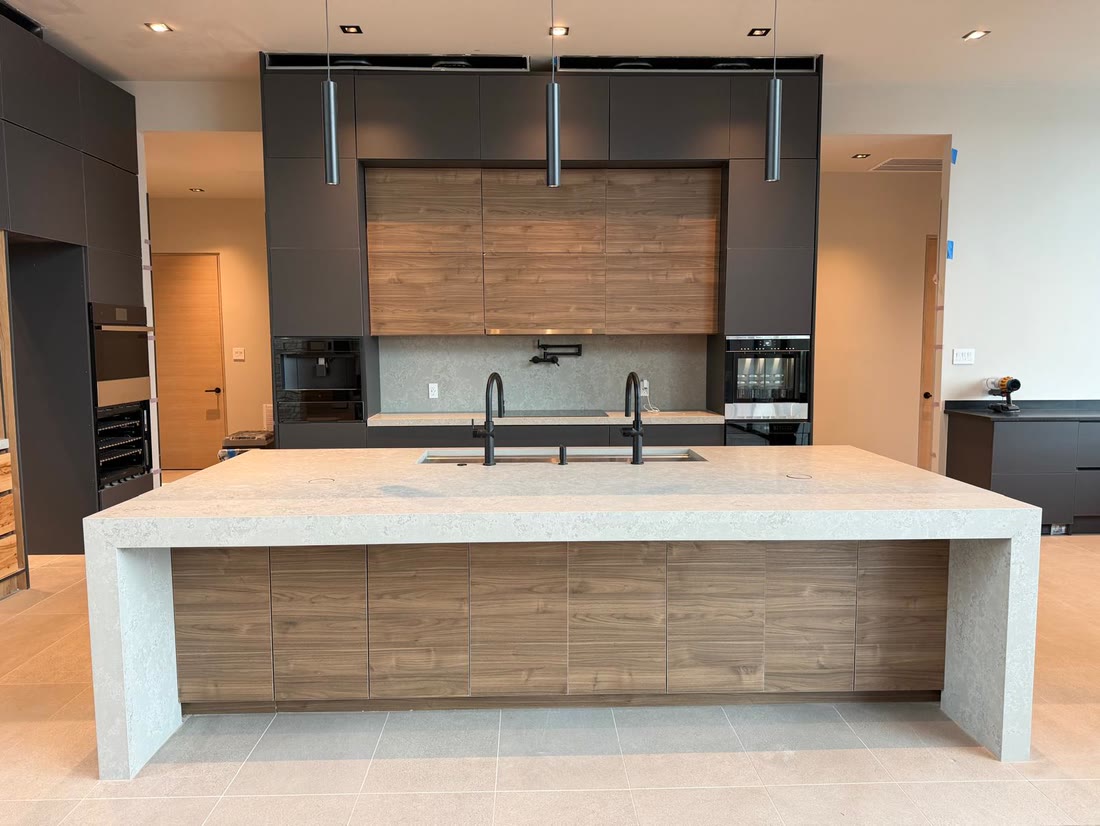 The kitchen island straight on, quartzite waterfall over walnut fronts, integrated coffee and wall ovens behind