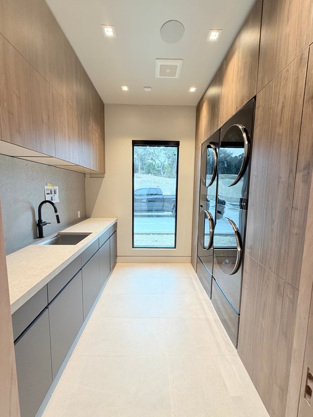 The scullery in pale timber and grey, with stacked machines and a window to the drive