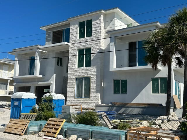 The house under construction from the side, stepped balconies and the stone centre, materials stacked in the foreground
