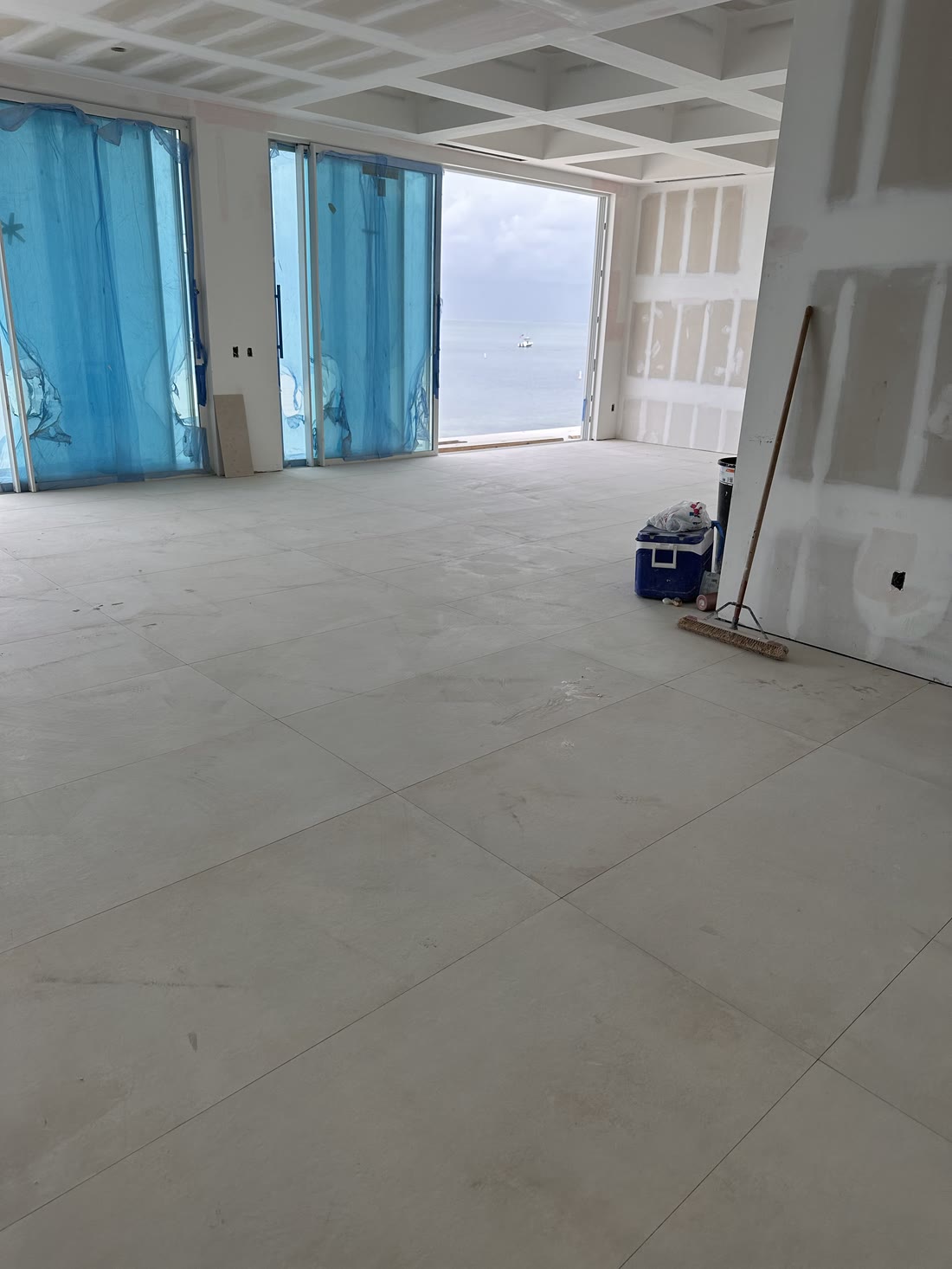 The main floor under construction: large format pale tile laid wall to wall, a coffered ceiling in, full height glazing openings taped in blue film, the sea beyond